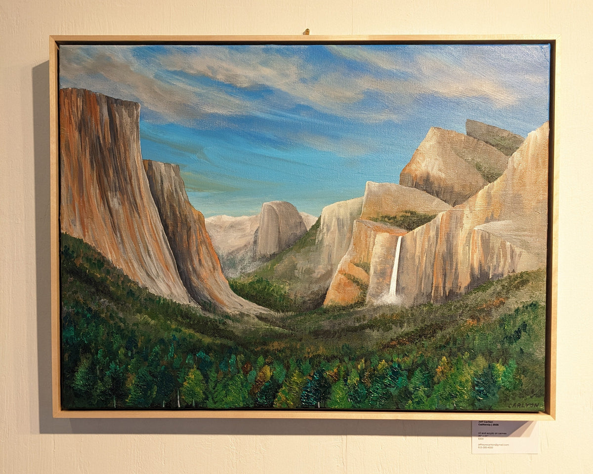 A framed landscape painting of a canyon valley with tall rock cliffs, a waterfall, green forest, and a blue sky, displayed on a beige wall.