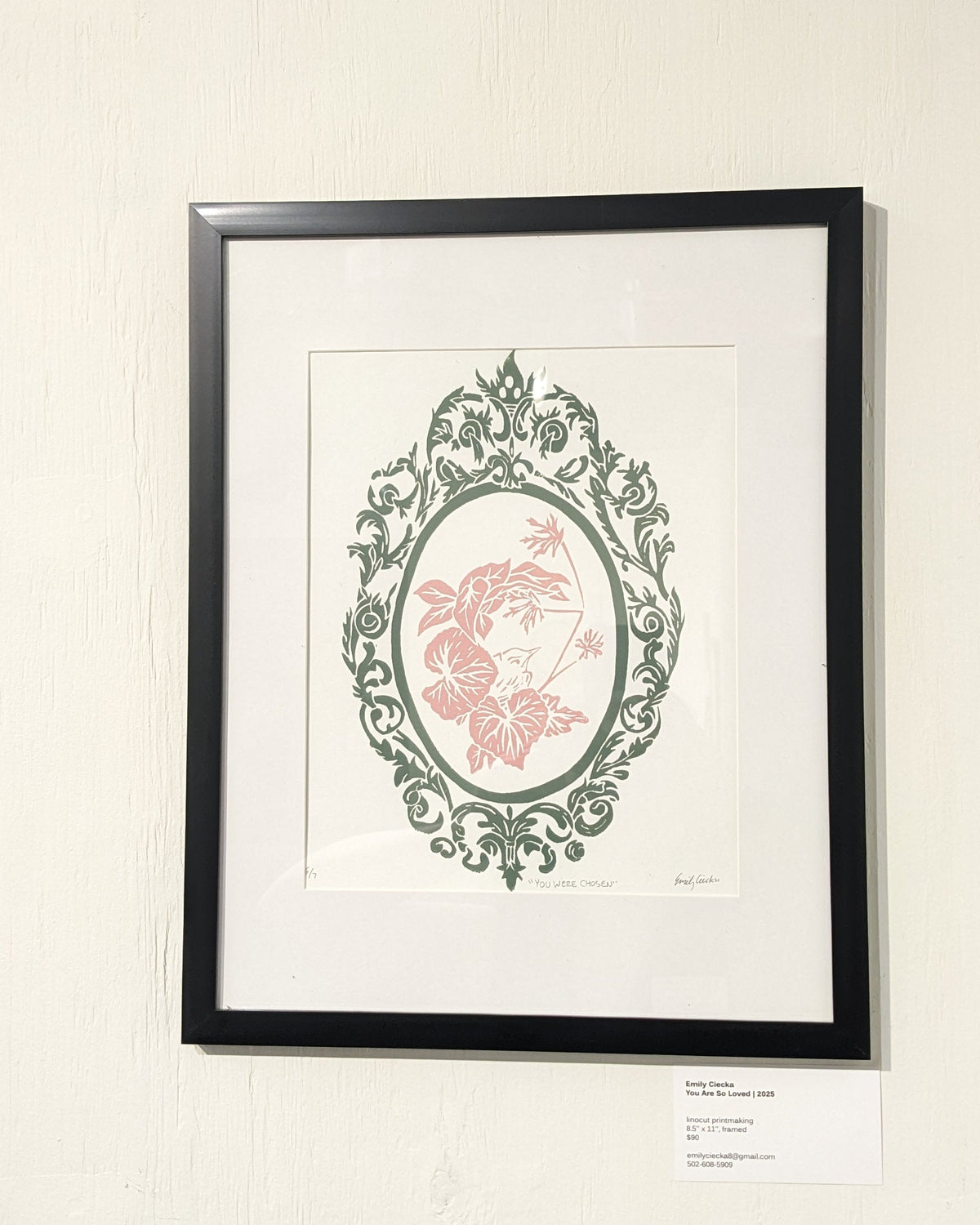 Framed artwork of pink bird surrounded by leaves with an ornate green design encircling them. 
