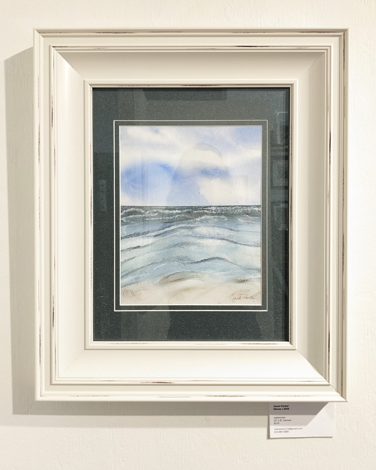 A framed watercolor painting of an ocean landscape with blue waves and a sandy beach under a cloudy sky, presented in a distressed white frame with a dark mat on a gallery wall.