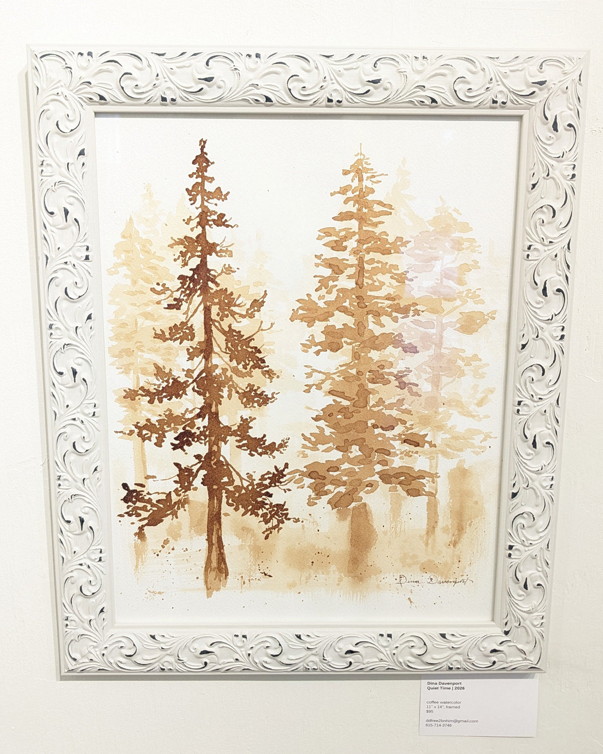 A framed watercolor painting of two tall pine trees in warm brown and beige tones, set in an ornate white carved frame and displayed on a light wall with small informational cards below.
