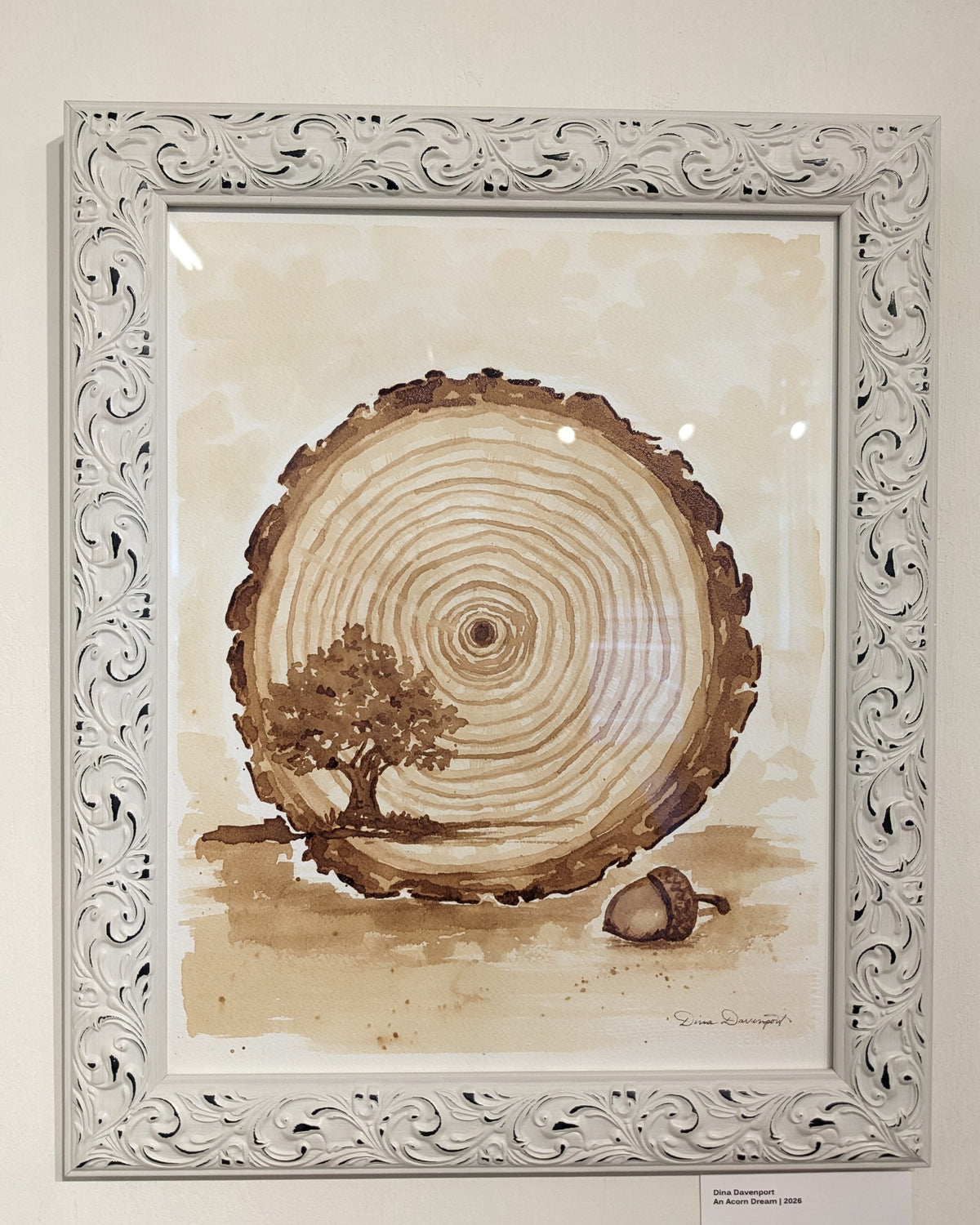 A framed art print featuring a sepia-toned watercolor illustration of a tree stump cross-section with a small tree and acorn, set in an ornate white carved frame on a beige wall.