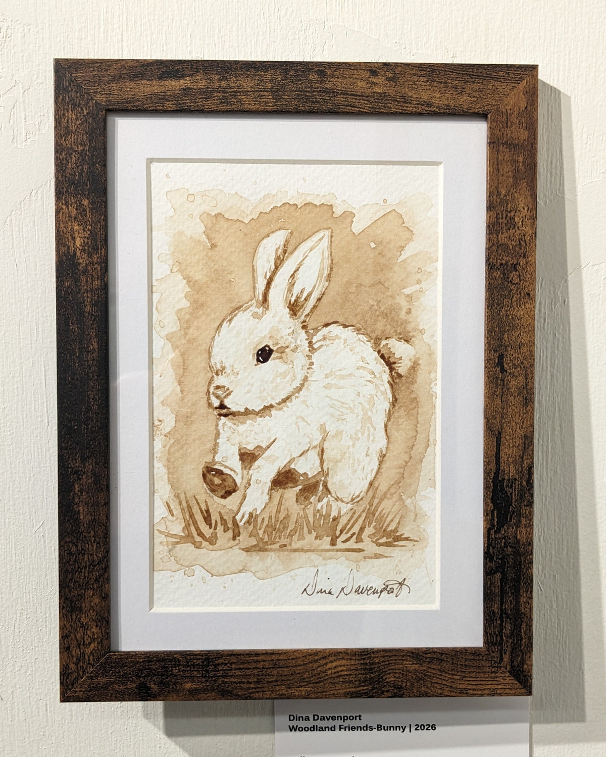 A framed art print featuring a sepia-toned drawing of a rabbit, mounted on a textured off-white wall with informational cards visible above and below the frame.
