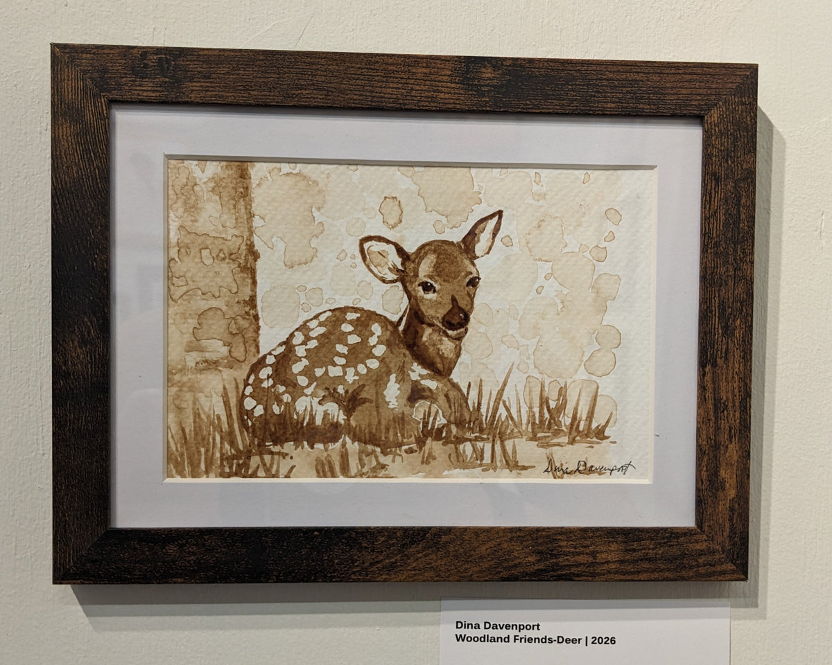 A framed watercolor painting of a woodland fawn, displayed on a gallery wall with informational placards. The artwork features a brown and beige fawn with white spots on a textured sepia background, enclosed in a dark brown wooden frame with a white mat.